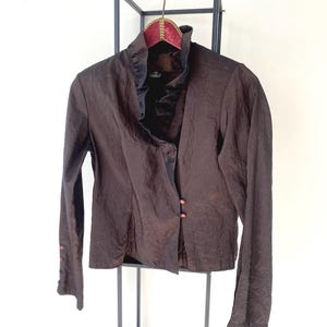 以下が含まれることがあります： フリル襟とボタン留めが1つ付いた、茶色の長袖ブラウス。ブラウスは黒い金属製のスタンドにかけられています。