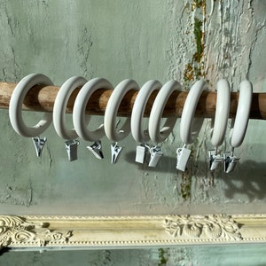 May include: White plastic curtain rings with silver clips. The rings are hanging on a wooden rod.