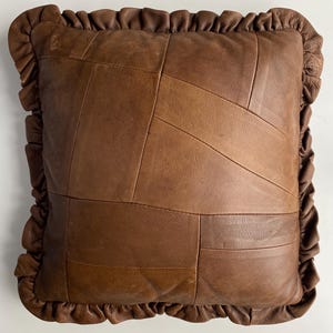 Pode incluir: Uma almofada quadrada de couro castanho com um design de patchwork. A almofada apresenta uma borda com babados e é feita de várias tonalidades de couro castanho. Um elemento decorativo para casa.