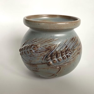 Può includere: Un piccolo vaso rotondo in ceramica grigio-blu tenue. Il vaso presenta un design a rilievo dettagliato di spighe di grano in tonalità marroni. Il bordo è marrone più scuro. Questo pezzo decorativo è adatto per l'arredamento della casa.
