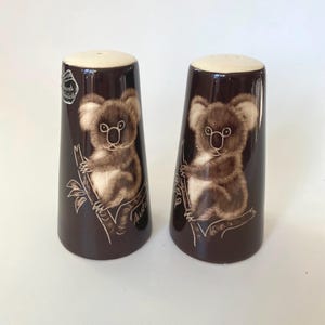 May include: A set of two brown ceramic salt and pepper shakers with a koala bear design. The shakers are decorated with a brown koala bear sitting on a tree branch. The text "Australia" is printed on the bottom of the shakers.