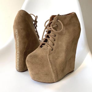 May include: Tan suede wedge ankle boots with lace-up closures. The boots have a platform sole and a stitched detail along the back. The wedge heel provides height and support, making them a stylish footwear option.