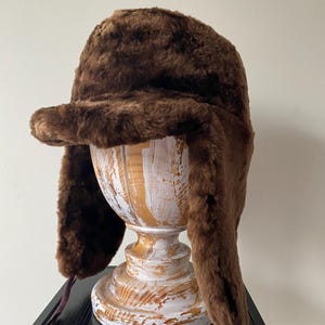 Pode incluir: Um chapéu de caçador de pele sintética castanha com abas para as orelhas e aba. O chapéu é exibido em um suporte de madeira decorativo. O chapéu tem uma aparência macia e texturizada e foi projetado para fornecer calor e cobertura.