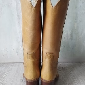 K&ouml;nnte beinhalten: Ein Paar hellbraune Leder-Cowboystiefel mit hohem Absatz und Rei&szlig;verschluss. Die Stiefel haben eine spitze Oberseite und einen braunen Absatz. Der Markenname ist auf der Seite der Stiefel sichtbar.