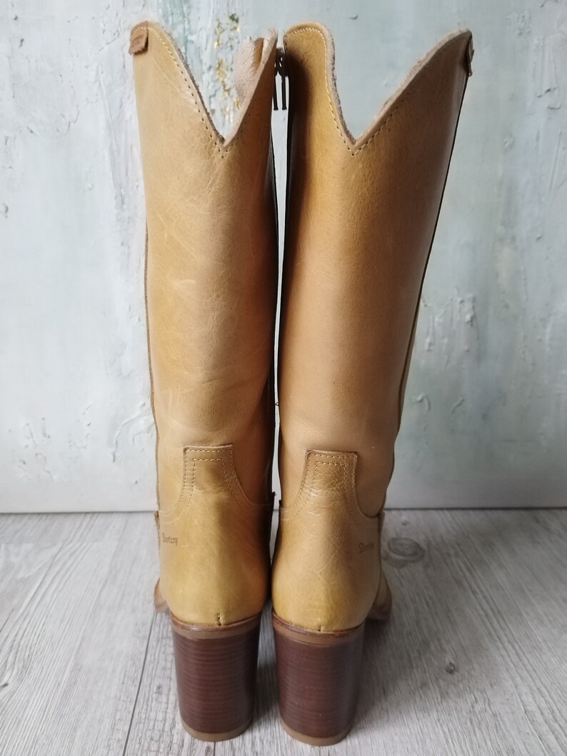 K&ouml;nnte beinhalten: Ein Paar hellbraune Leder-Cowboystiefel mit hohem Absatz und Rei&szlig;verschluss. Die Stiefel haben eine spitze Oberseite und einen braunen Absatz. Der Markenname ist auf der Seite der Stiefel sichtbar.