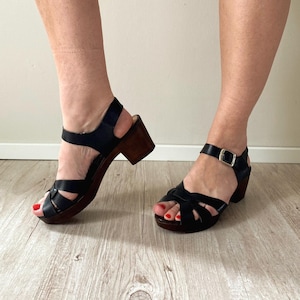 May include: A pair of black leather sandals with a woven strap design across the toe, a buckled ankle strap, and a dark brown wooden block heel. The sandals feature a sturdy construction with a rounded toe opening and a metallic silver buckle.