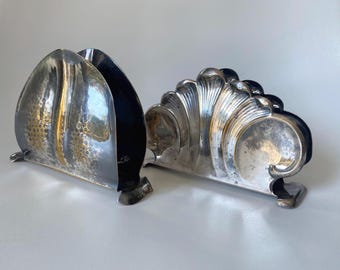 Peut inclure: Deux porte-serviettes en métal argenté. Celui de gauche présente une forme arquée lisse avec un motif texturé en nid d'abeille. Celui de droite arbore un design en forme de coquille festonnée avec des stries verticales et une base courbée.