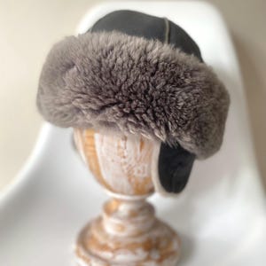 Könnte beinhalten: Eine dunkelgraue Wintermütze mit einem dicken, flauschigen hellbraunen Kunstfellbesatz am Rand und an den Ohrenklappen. Die Krone der Mütze besteht aus glatten, dunkelgrauen Stoffbahnen mit sichtbaren Nähten für eine strukturierte Form.