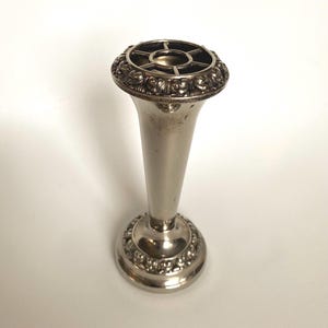 May include: A silver-toned bud vase with a flared top and a round base. The top features a grid design, and the base and top rim are adorned with a floral pattern. The vase is approximately 15 cm tall.