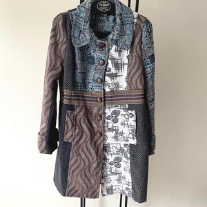 Puede incluir: Un abrigo de manga larga con un diseño de patchwork de varios tejidos en tonos marrón, azul, blanco y negro. El abrigo tiene un cuello ancho, cierre frontal con botones marrones y una banda horizontal con motivos decorativos en la cintura.