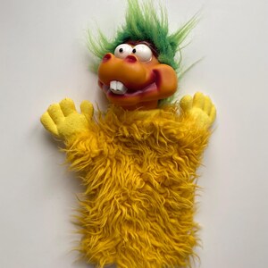 Op de afbeelding: Een gele, pluche pop met een groene mohawk en een brede glimlach. De pop heeft grote, ronde ogen en een feloranje mond.