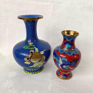 Pode incluir: Dois vasos decorativos cloisonné. O vaso maior é azul com um desenho de peixe dourado e flor de lótus. O vaso menor é vermelho com um desenho de dragão azul e branco. Ambos os vasos têm bordas douradas.
