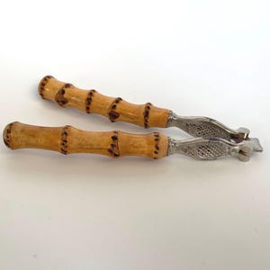 Puede incluir: Un par de cascanueces de bambú con un mecanismo de cascanueces de metal plateado. Los mangos son de bambú natural de color marrón y beige.