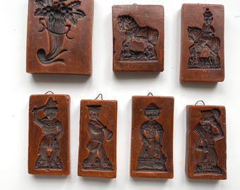 Peut inclure: Un ensemble de sept moules à biscuits rectangulaires en bois brun, chacun présentant un motif en relief sculpté distinct, notamment une corne d'abondance, un cheval, un cavalier et divers personnages, avec des boucles métalliques au sommet.