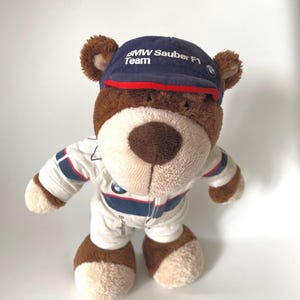 May include: A brown plush teddy bear wearing a white racing jumpsuit with blue and red stripes and a navy blue baseball cap featuring the white text BMW Sauber F1 Team and a small circular logo on the side of the cap brim.