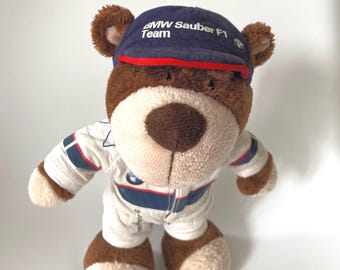 Peut inclure: Un ours en peluche marron portant une combinaison de course blanche avec des rayures bleues et rouges et une casquette bleu marine avec le texte blanc BMW Sauber F1 Team et un petit logo circulaire sur le côté de la visière.