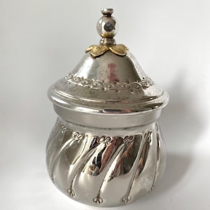 May include: A silver-toned lidded jar with a reflective finish, featuring a rounded body with vertical swirling indentations. The lid is topped with a small spherical finial and a gold-colored floral accent, with a decorative embossed border.