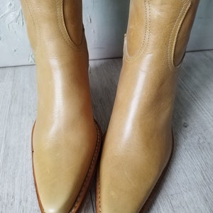 K&ouml;nnte beinhalten: Ein Paar hellbraune Leder-Cowboystiefel. Die Stiefel haben spitze Zehen, dekorative N&auml;hte und einen mittelhohen Absatz. Das Leder hat eine glatte Oberfl&auml;che und die Stiefel scheinen neu oder in ausgezeichnetem Zustand zu sein. Die Stiefel sind neutral gef&auml;rbt.