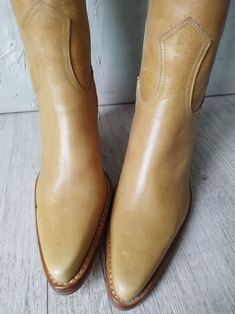 K&ouml;nnte beinhalten: Ein Paar hellbraune Leder-Cowboystiefel. Die Stiefel haben spitze Zehen, dekorative N&auml;hte und einen mittelhohen Absatz. Das Leder hat eine glatte Oberfl&auml;che und die Stiefel scheinen neu oder in ausgezeichnetem Zustand zu sein. Die Stiefel sind neutral gef&auml;rbt.