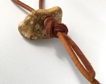 Peut inclure: Un collier composé d'un pendentif en pierre de couleur beige, à la texture irrégulière et parsemé de petites marques circulaires blanches. La pierre est suspendue à un fin cordon en cuir marron noué solidement autour du centre.