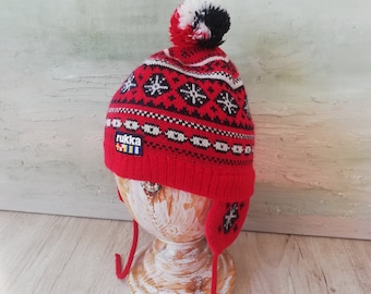 May include: Red knitted winter hat with a white and black pom pom on top. The hat has a black and white snowflake pattern and the word "Rukka" is embroidered on the side.