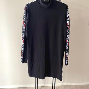 Puede incluir: Un vestido negro de manga larga con cuello alto que presenta una banda de marca en blanco, rojo y negro a lo largo de cada manga. La prenda est&aacute; confeccionada en tela negra lisa e incluye el logotipo de FILA repetido en las franjas laterales.