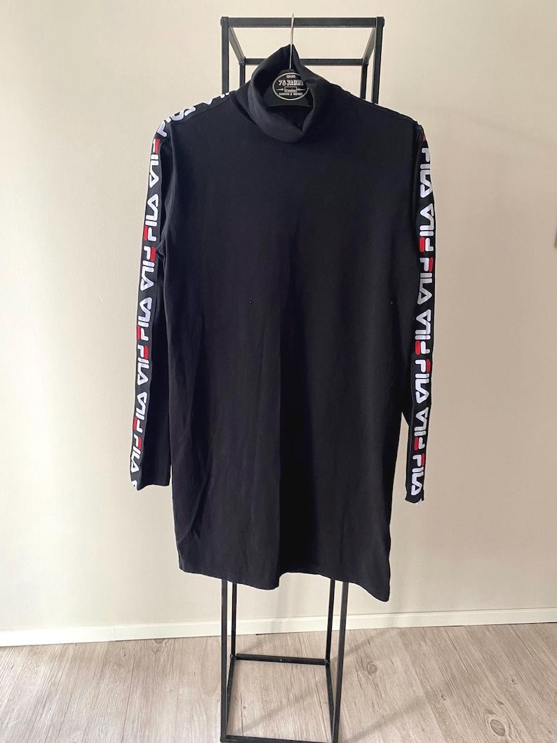 Puede incluir: Un vestido negro de manga larga con cuello alto que presenta una banda de marca en blanco, rojo y negro a lo largo de cada manga. La prenda est&aacute; confeccionada en tela negra lisa e incluye el logotipo de FILA repetido en las franjas laterales.