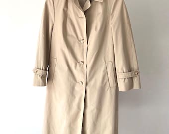 May include: A tan trench coat with a classic design, featuring a collared neckline, button-down front, and long sleeves. The coat has a tailored fit, pockets, and buttoned cuffs. It is hanging on a black metal rack.