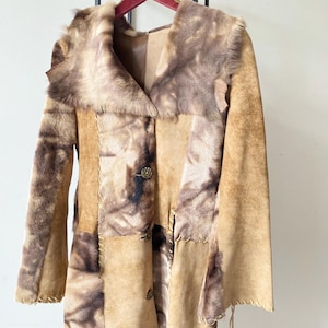 Op de afbeelding: Een lange, bruine en beige patchwork suède jas met een bontkraag. De jas heeft een strik in de taille en een knoopsluiting.