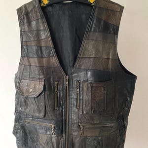 Op de afbeelding: Een vintage leren vest in bruin- en zwarttinten, met een patchwork design. Het vest heeft meerdere zakken met ritssluiting en een zak met knoop. De voorkant heeft een volledige ritssluiting. Het vest hangt aan een zwarte hanger.
