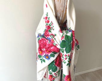 Peut inclure: Un châle de couleur crème orné d'un motif floral vibrant composé de grandes roses roses, de petites fleurs bleues et de feuilles vertes. Le tissu est drapé sur un mannequin et terminé par de longues franges blanches fines sur les bords.