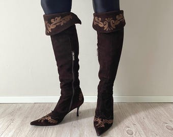 Peut inclure: Bottes hautes en daim marron foncé avec un bout pointu, un talon aiguille fin et une fermeture éclair latérale. Les bottes sont ornées de broderies cuivrées complexes sur le revers replié et sur la zone du bout pointu.