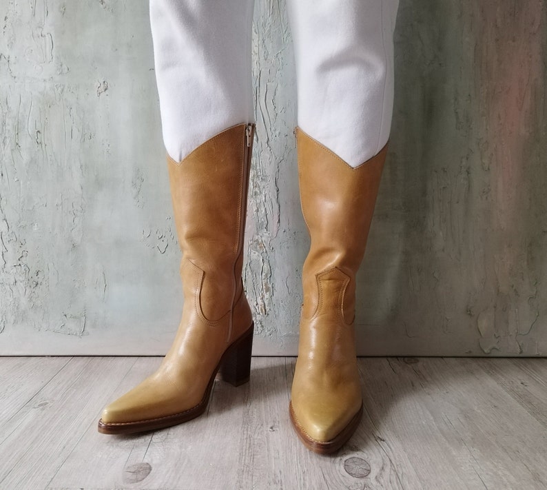 K&ouml;nnte beinhalten: Hellbraune Cowboy-Stiefel aus Leder mit spitzer Zehenpartie und hohen Blockabs&auml;tzen. Die kniehohen Stiefel haben einen Rei&szlig;verschluss hinten und ein V-f&ouml;rmiges Detail oben. Die Abs&auml;tze sind etwa 7,6 cm hoch.