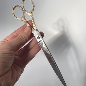 May include: A pair of scissors featuring long, pointed silver-toned metal blades and ornate gold-toned handles. The handles are decorated with intricate floral and scrollwork engravings, and a central screw connects the two blades together.