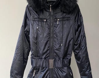 Peut inclure: Une combinaison de neige noire dotée d'un col montant, d'une fermeture éclair intégrale sur le devant et d'une capuche bordée de fausse fourrure noire. Le vêtement comprend une large ceinture élastique avec une boucle en métal argenté.