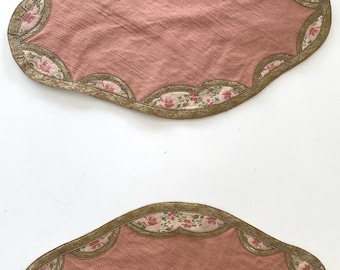 Peut inclure: Deux sets de table ovales en tissu avec un panneau central rose poudré uni. La bordure présente un contour festonné orné d'un motif floral de roses roses et de feuilles vertes sur fond crème, le tout fini par un galon doré métallique.