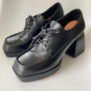 May include: A pair of black lace-up shoes featuring a square toe, a thick platform sole, and a chunky block heel. The shoes have a smooth finish, matching black laces, and a tan-colored interior lining visible at the heel opening.
