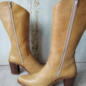 K&ouml;nnte beinhalten: Ein Paar hellbraune Lederstiefel mit quadratischer Spitze und Blockabsatz. Die Stiefel haben einen seitlichen Rei&szlig;verschluss und einen hellfarbigen Rei&szlig;verschluss. Der Absatz ist ca. 7,6 cm hoch.