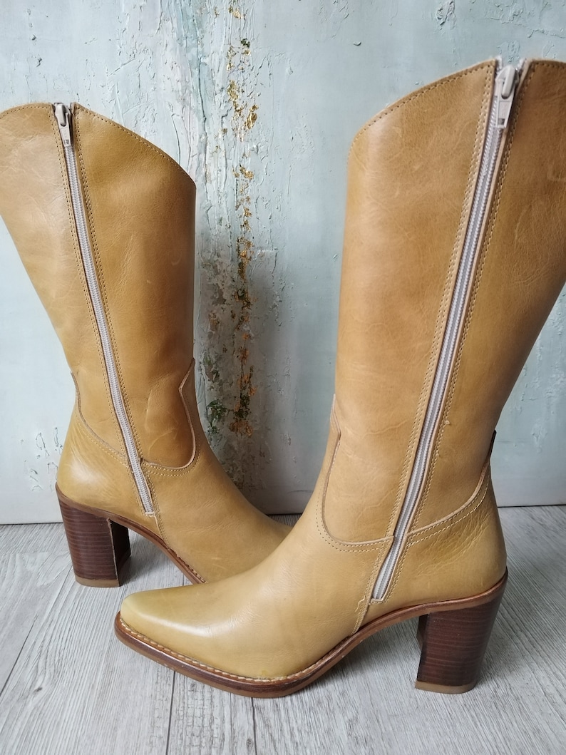 K&ouml;nnte beinhalten: Ein Paar hellbraune Lederstiefel mit quadratischer Spitze und Blockabsatz. Die Stiefel haben einen seitlichen Rei&szlig;verschluss und einen hellfarbigen Rei&szlig;verschluss. Der Absatz ist ca. 7,6 cm hoch.