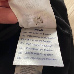 Puede incluir: Una etiqueta de cuidado de tela blanca unida a un textil negro. La etiqueta presenta la marca Fila y enumera la composici&oacute;n del material como 95% algod&oacute;n y 5% elastano en varios idiomas, incluidos ingl&eacute;s, alem&aacute;n, franc&eacute;s, italiano, holand&eacute;s y espa&ntilde;ol.