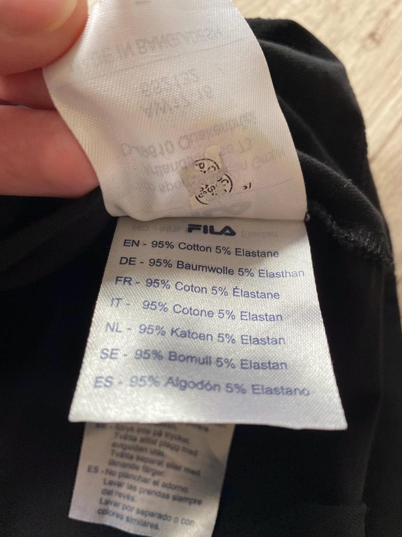 Puede incluir: Una etiqueta de cuidado de tela blanca unida a un textil negro. La etiqueta presenta la marca Fila y enumera la composici&oacute;n del material como 95% algod&oacute;n y 5% elastano en varios idiomas, incluidos ingl&eacute;s, alem&aacute;n, franc&eacute;s, italiano, holand&eacute;s y espa&ntilde;ol.