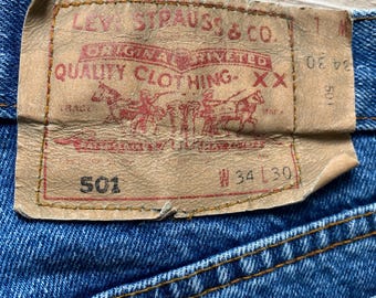 Peut inclure: Jean en denim bleu avec une étiquette en cuir beige sur la ceinture arrière. L'étiquette porte le texte Levi Strauss & Co. Original Riveted Quality Clothing XX, le numéro 501 et la taille W 34 L 30 avec des coutures contrastées orange.