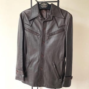 Pode incluir: Casaco de couro castanho com fecho de botões, dois bolsos no peito e dois bolsos laterais com zíper. O casaco tem uma gola larga e punhos abotoados. Está pendurado num suporte de metal preto.