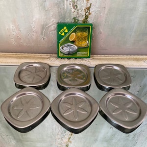 May include: Six silver metal coasters with a flower design. The coasters are square and have a raised edge. The coasters are packaged in a green box with the word "Untersetzer" printed on it.