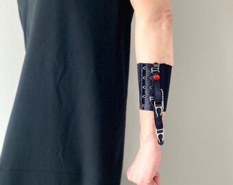 Peut inclure: Une manchette en tissu noir dotée d'un laçage de style corset avec des œillets métalliques, un ornement en forme de pierre précieuse ovale rouge et une sangle noire pendante avec une attache en métal argenté, portée sur l'avant-bras.