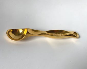 Peut inclure: Une cuillère en métal doré poli avec un bol arrondi et un manche profilé. Le manche présente une indentation allongée et un petit trou circulaire à l'extrémité pour la suspension. La surface est réfléchissante et lisse avec une finition métallique.