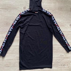 Puede incluir: Una camiseta negra de manga larga con cuello alto que presenta el logotipo de Fila en el pecho y una cinta con el logotipo de Fila repetido en blanco y rojo a lo largo de ambas mangas. La prenda se muestra extendida sobre madera clara.