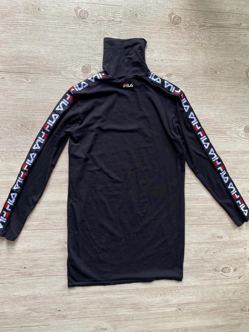 Puede incluir: Una camiseta negra de manga larga con cuello alto que presenta el logotipo de Fila en el pecho y una cinta con el logotipo de Fila repetido en blanco y rojo a lo largo de ambas mangas. La prenda se muestra extendida sobre madera clara.