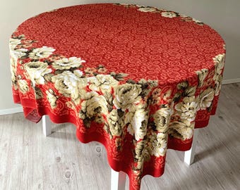 Peut inclure: Une nappe ronde présentant un fond rouge vif avec un motif répétitif complexe et une large bordure de grandes roses couleur crème avec des feuilles vert foncé drapées sur les bords d'une table blanche.