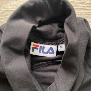 Puede incluir: Una camiseta negra de cuello alto con una etiqueta interior blanca con el logotipo de FILA en rojo y azul y una etiqueta separada que indica la talla M. La prenda presenta detalles de rayas rojas, blancas y azules en las costuras de los hombros.